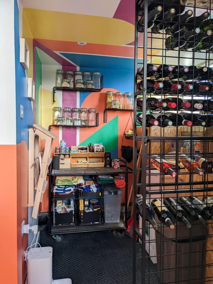 large walk in pantry with walls painted in yellow, orange, blue, green, purple shapes, storage cart and shelves with storage canisters on far wall, small ladder mounted on left wall, floor to ceiling wine rack in foreground, more storage behind