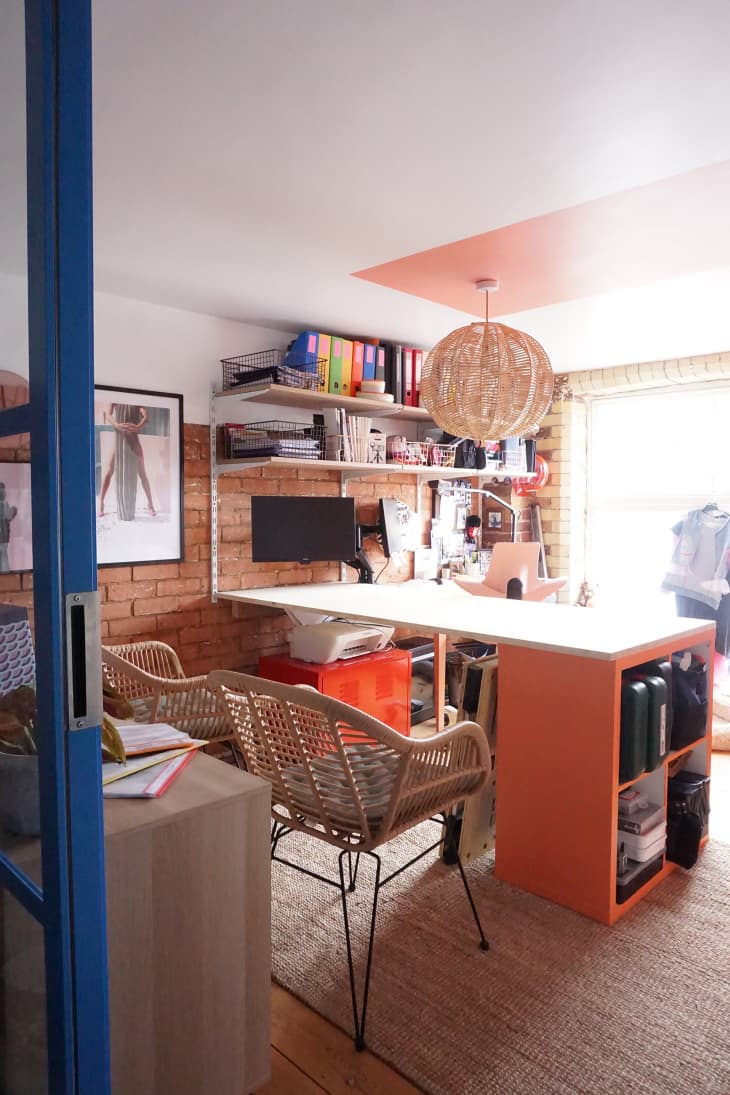 Study/home office. Orange and white desk, 2 cane chairs, rattan globe pendant light, exposed brick wall, blue paned window/door, neutral rug, coral accents painted on ceiling and wall