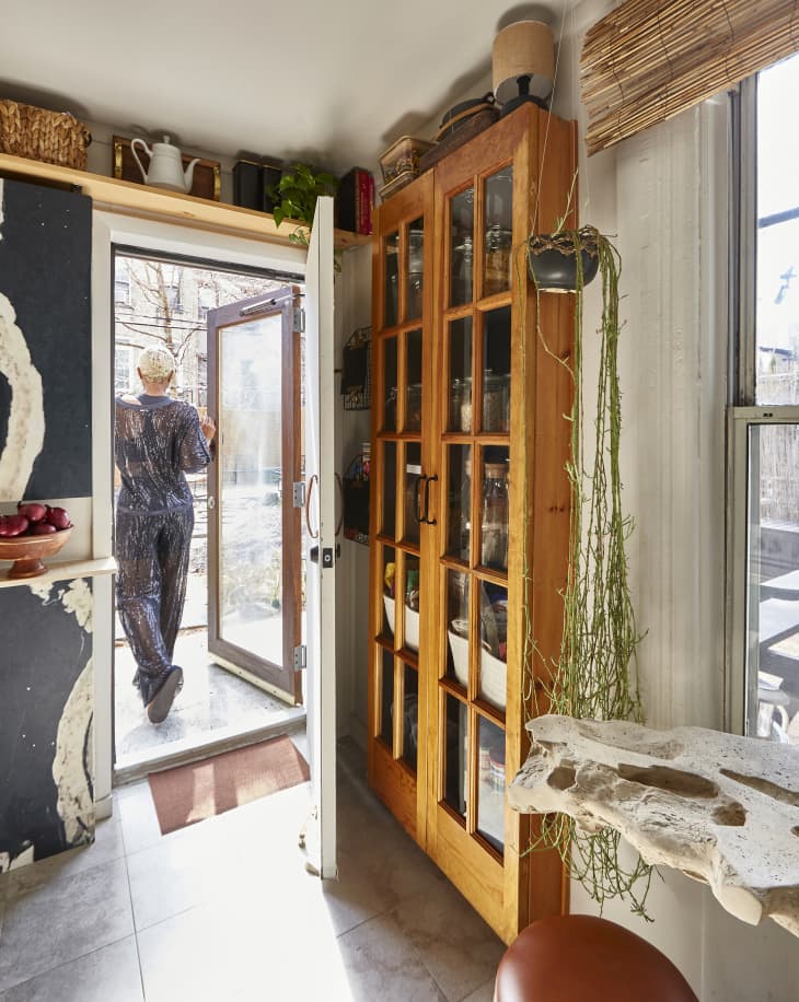 View of someone walking out the door of their kitchen to the outside. Kitchen has a tall wood storage hutch, and a ceiling shelf with teapots, baskets. You can see a hint of a black and white painted cabinet