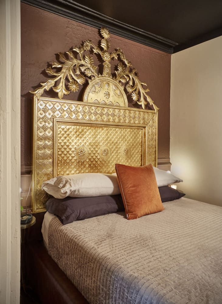 bedroom with brown accent wall over bed. Bed has ornate gold headboard, warm and neutral colored bedding