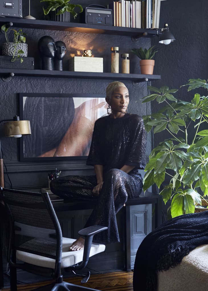 Kim sitting on her desk in workspace. Dark blue walls, lots of art objects on shelves above, large tree/plant