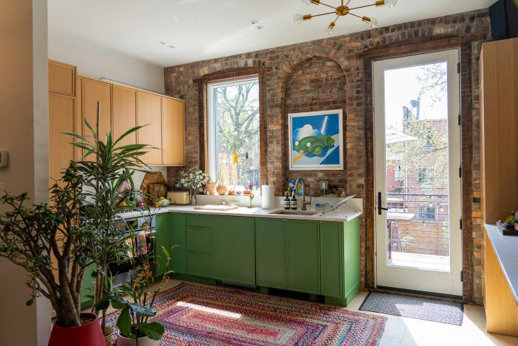 Light filled, brick walled kitchen with green lower cabinets and wooden upper cabinets.