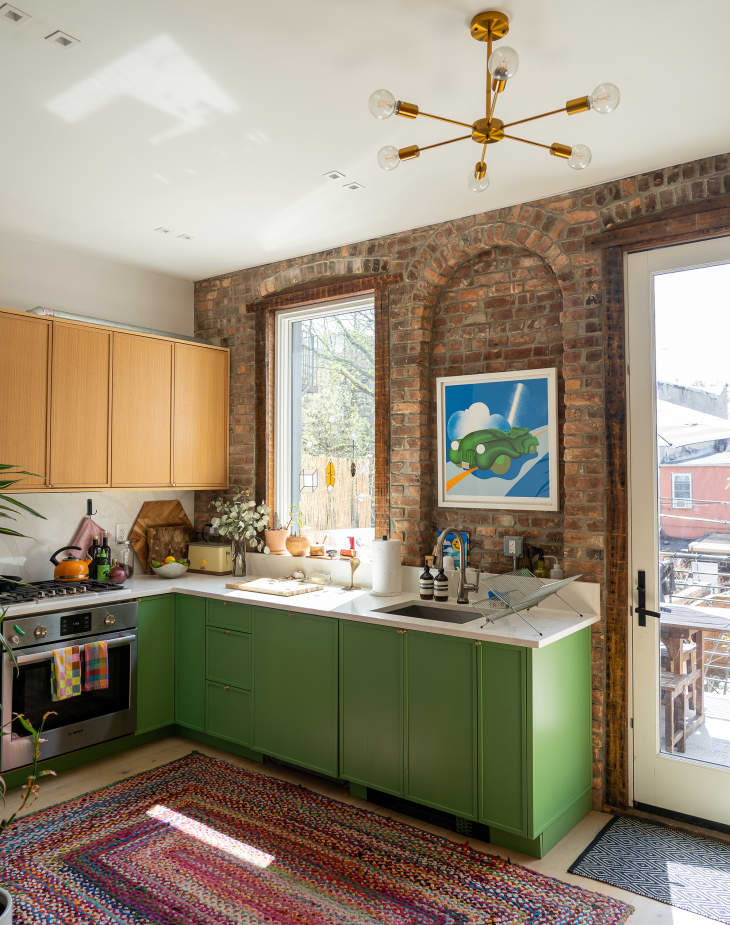 Green lower cabinets in brick walled kitchen with multi-armed pendent light on ceiling.