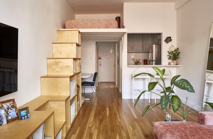 View from living room into loft space. White walls, wooden shelves that act as steps to the loft, wood floors, large plant. Kitchen also in view in background