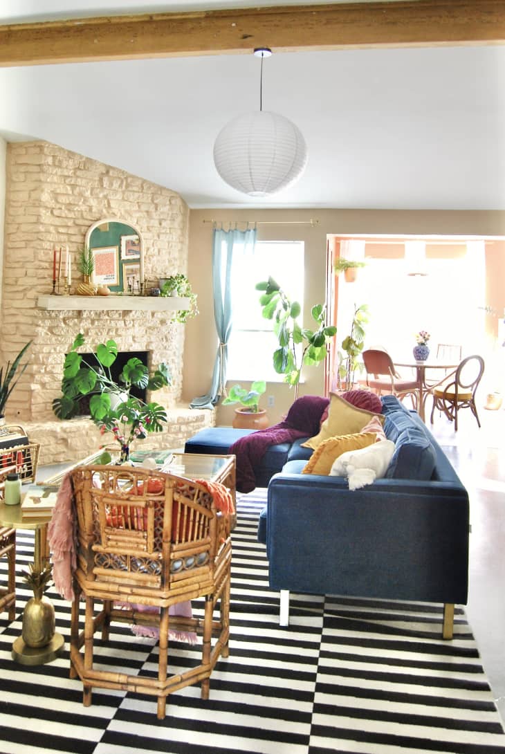 Living room: pale brick fireplace, potted plants, mantel with arch shaped mirror, candles, objets, black and white striped area rug, blue velvet sofa. View into sunny dining room behind