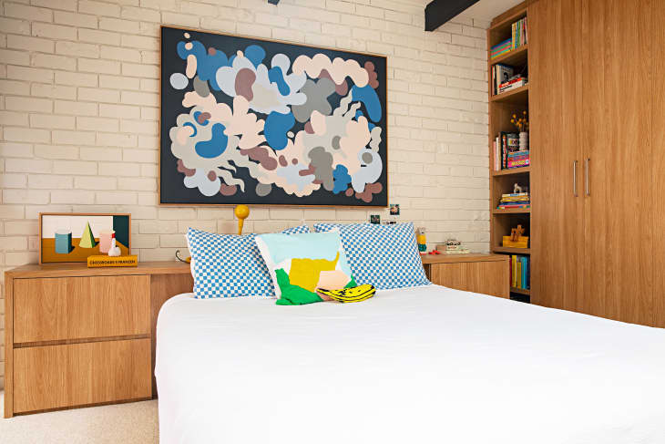 bedroom with painted white brick wall behind bed, white bed linens, Large wood closet and shelves on one wall, wood headboard with built-in side tables, art and throw pillows with blue accents