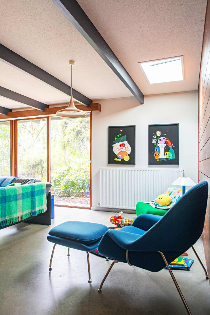 Part of a living space with large windows, exposed black beams, one wood paneled wall, blue accent chair, small kids green armchair, toys around. You can see the back of a sofa