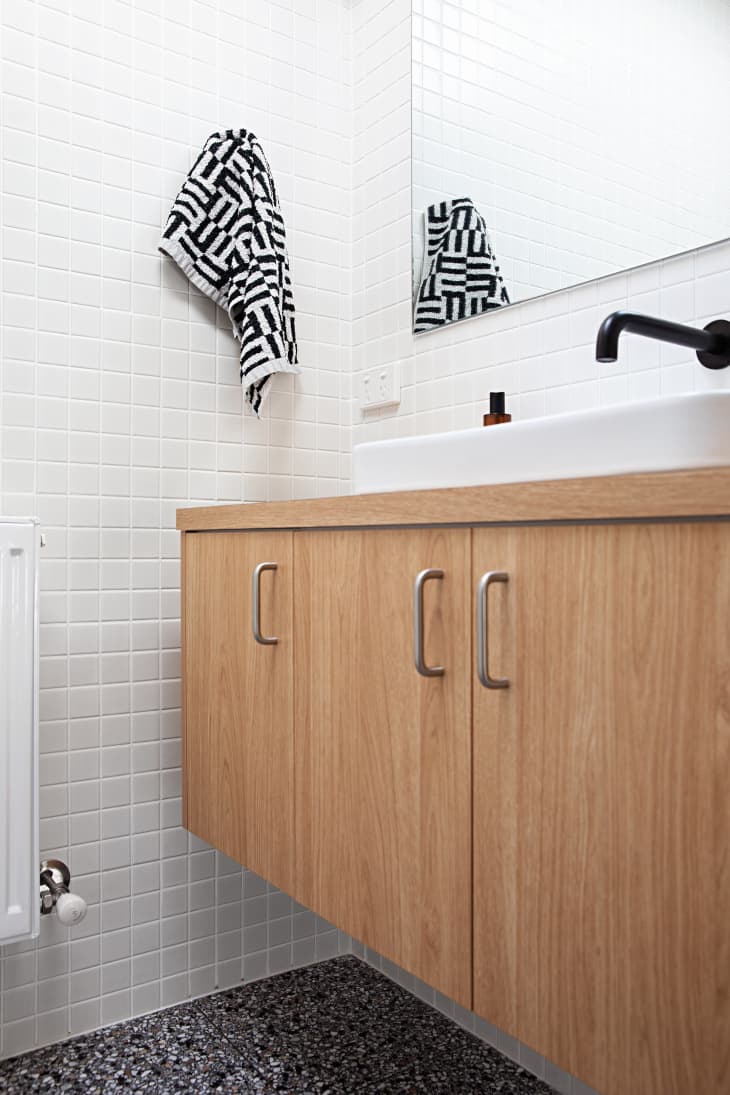 bathroom with white tile walls, black and white patterned hand towel, white sink with black hardware, wood cabinets