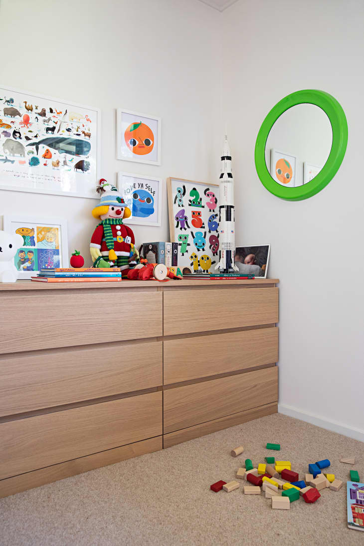 Detail of wood dresser in kids room. White walls, round green mirror, lots of toys and art