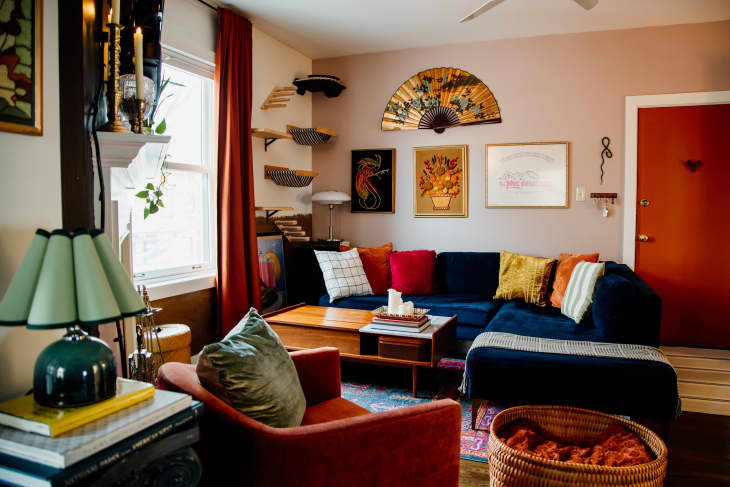living room with navy blue velvet sofa, red and gold throw pillows, rust colored velvet accent chair, large gold/floral fan over sofa as art, cat ladders and sling beds going up the wall, floor to ceiling rust curtains, rust/terracotta colored door