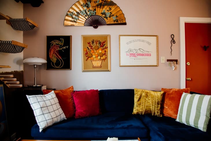 living room with navy blue velvet sofa, red and gold throw pillows, rust colored velvet accent chair, large gold/floral fan over sofa as art, cat ladders and sling beds going up the wall, floor to ceiling rust curtains, rust/terracotta colored door