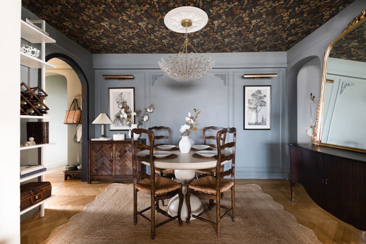 Dining room with wood floor, jute rug, dark wood sideboards, open shelving, blue-gray walls, chandelier and wallpapered ceiling.