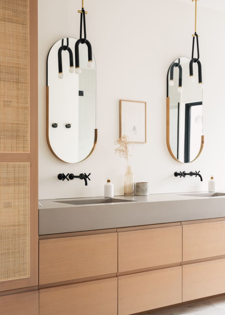 bathroom counter with 2 oval mirrors, black hanging light fixtures, white walls, wood drawers