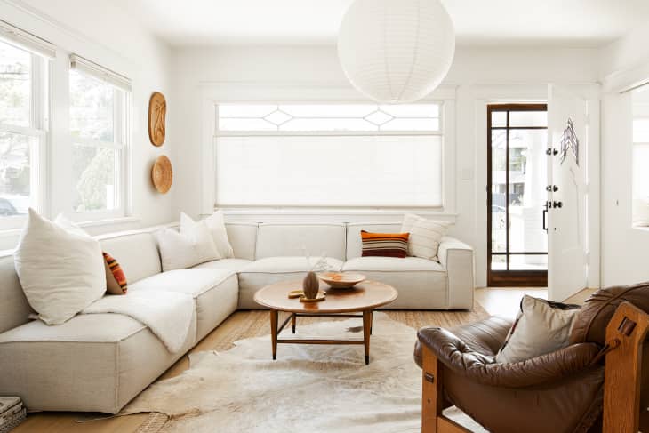 living room with large off white sectional sofa, brown leather armchair, white walls, large white round hanging lantern, hide area rug, large windows