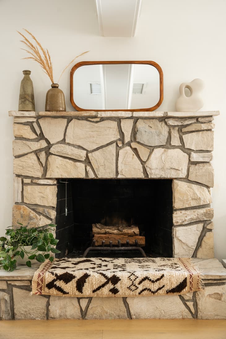 stone fireplace with neutral colors, mirror on mantel, vase and art objects, plant and rug on hearth
