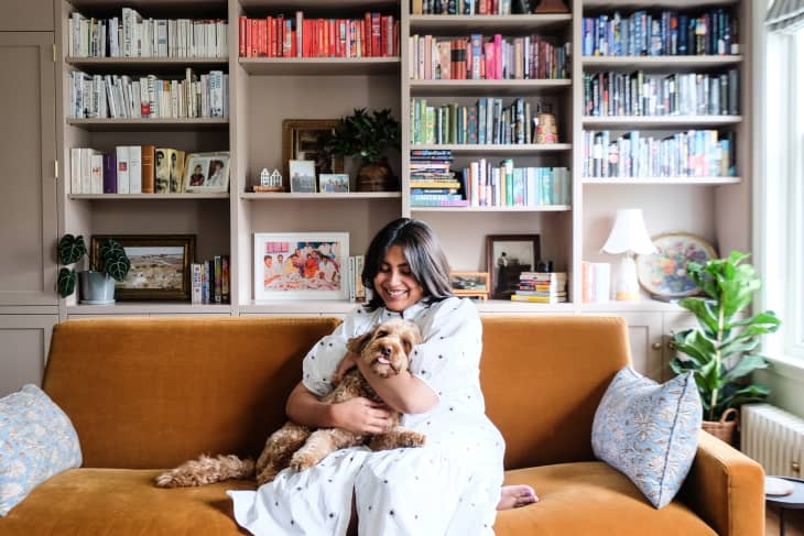 Dweller sits hugs dog on velvet ochre sofa in book filled living room.