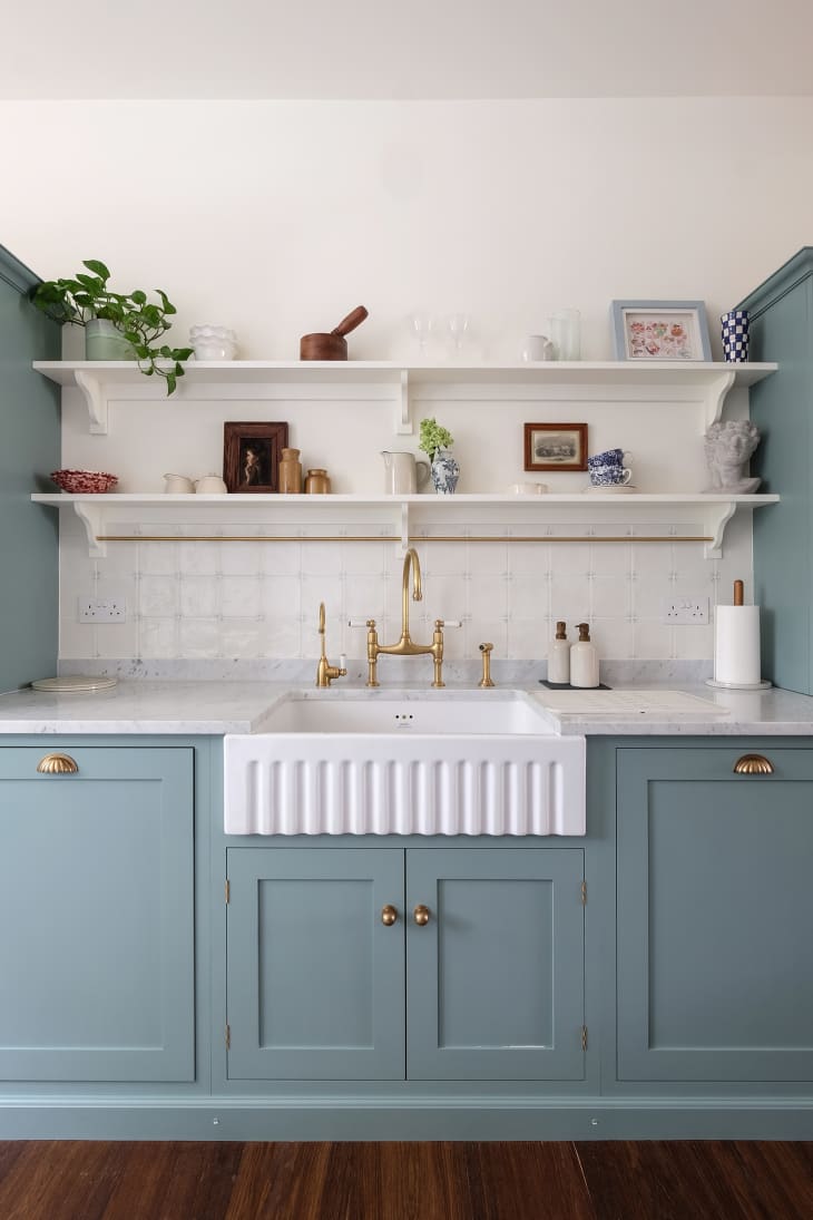 Decorative objects line open shelving in newly renovated kitchen with white and grey marbled surface and large white farm sink.
