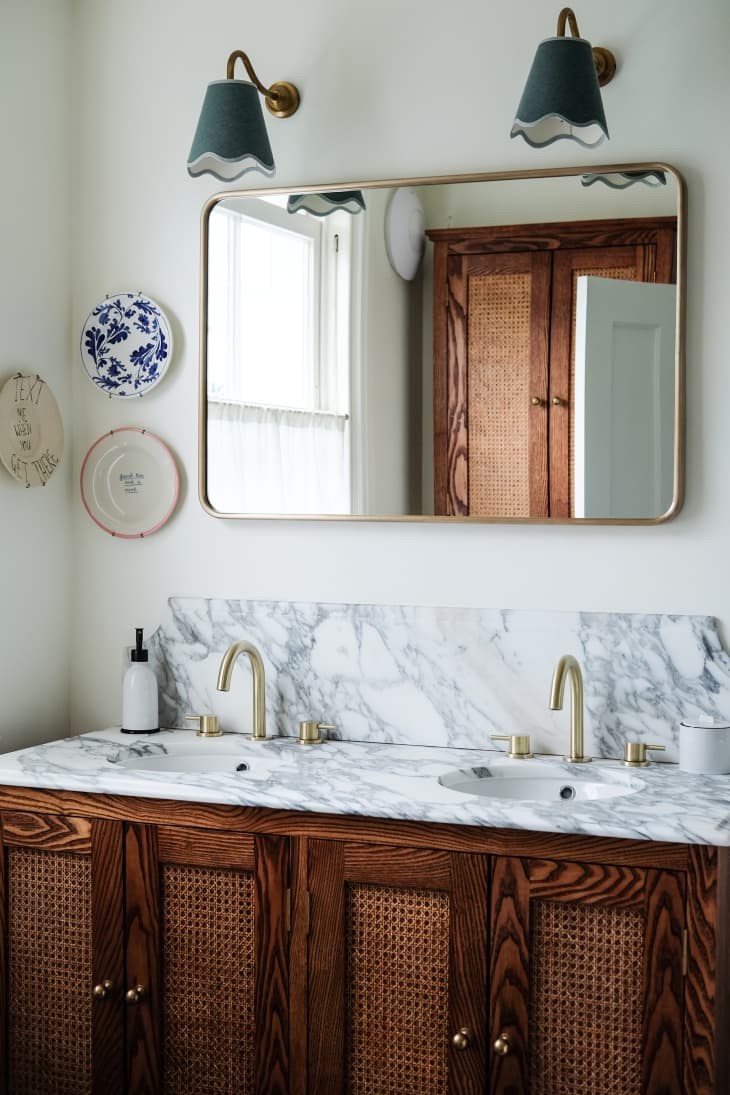 Green sconces mounted above gold framed rectangular mirror in newly renovated bathroom with gray and white marbled surface on caned wood bathroom vanity. Small plates displayed on wall beside bathroom vanity.