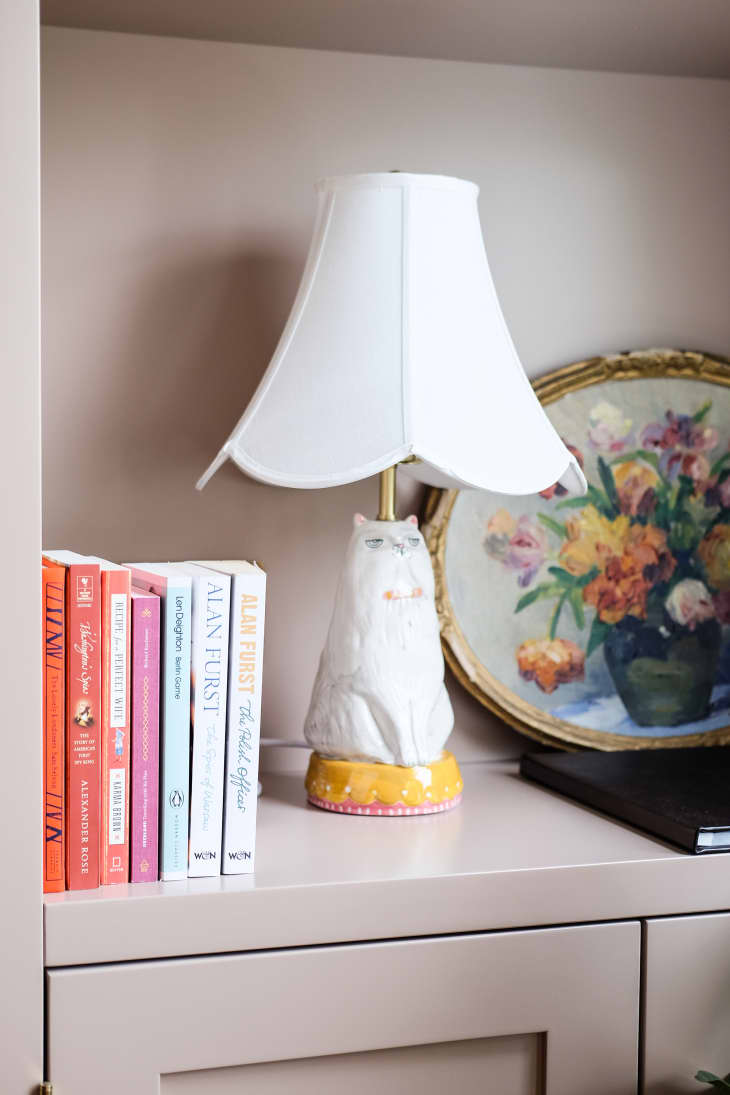 Lamp base with cat figurine and white lamp shade sits in bookshelf with books and small floral painting beside.