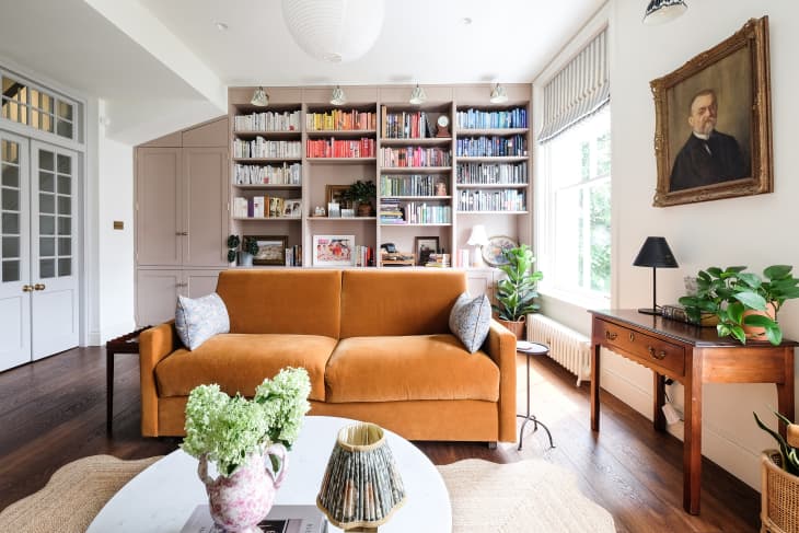 Ochre velvet sofa in book filled living room with vintage painted portrait mounted above wooden side table.