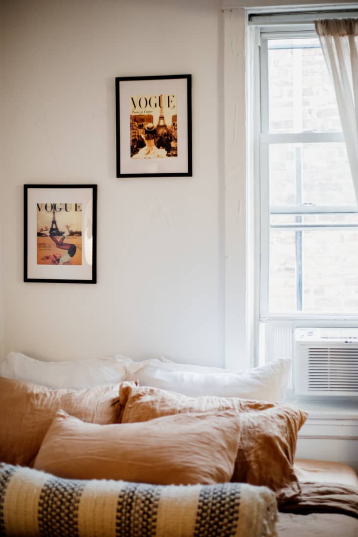 view into bedroom with white walls, bed with warm-colored linens and pillows, 2 black framed vintage vogue covers on wall, window with sheer curtains
