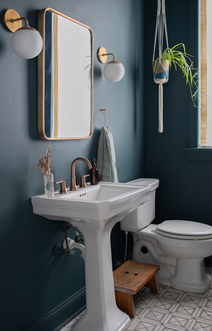 Bathroom with blue walls, white sink, brass accents, hanging plant, floor tiles with square pattern, small wood stool