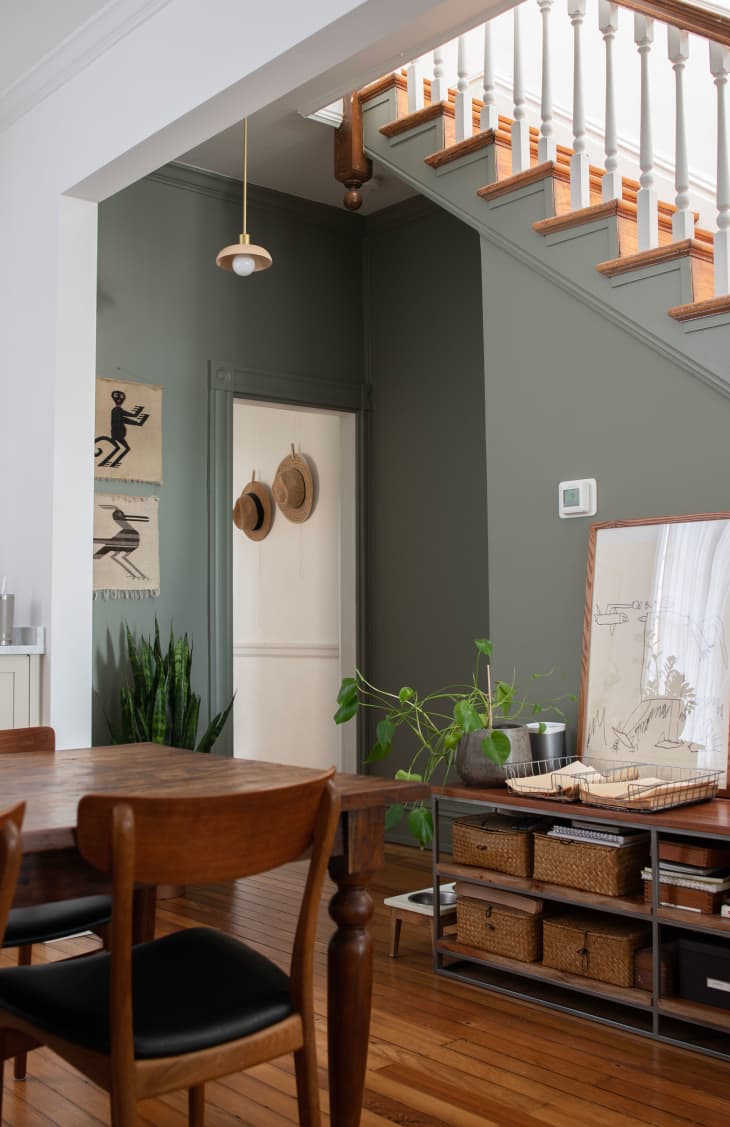 dining room with green walls, wood floors, wood dining table, wood and black midcentury chairs, shelves with organizing baskets, mirror and plant on top, Stairs leading up