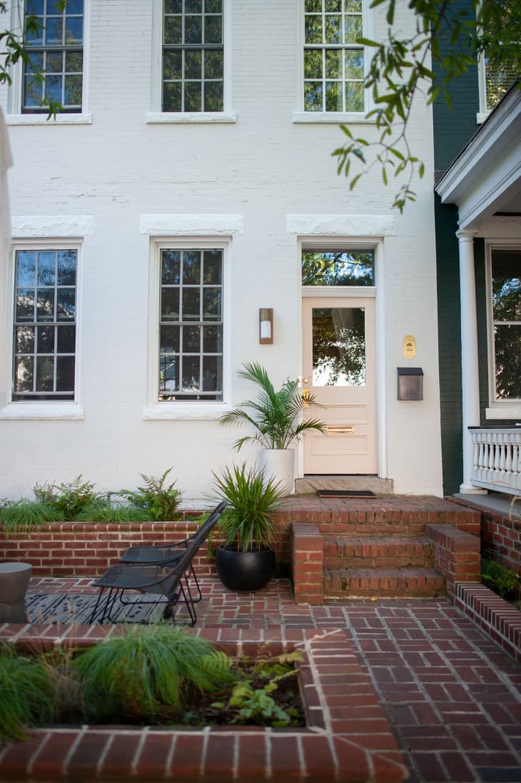 Outside of white building with brick stoop, stairs, front patio. Plants and 2 black chairs