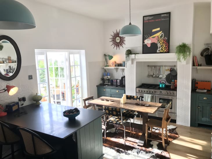 view looking down into kitchen/dining area. wood floor, wood dining table with mixed chairs, white walls, french doors with paned windows, deep green/blue cabinets and drawers, light teal half dome pendant lights, deep green blue island with 2 rattan and black chairs