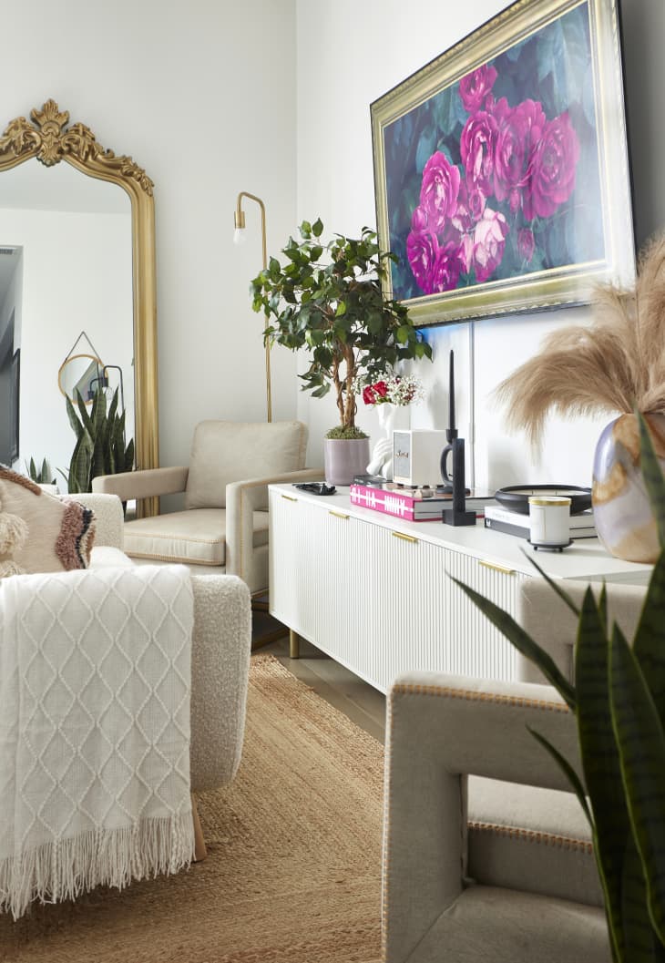 living space with neutral/pink cozy chairs, white walls, white credenza with gold pulls, large painting of flowers in gold frame, plants, gold, black, white and neutral accents, Gold ornate mirror on back wall with another neutral toned armchair