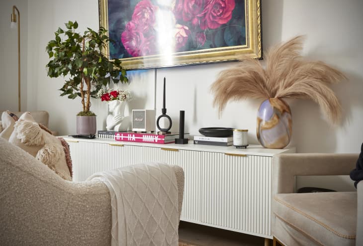 living space with neutral/pink cozy chairs, white walls, white credenza with gold pulls, large painting of flowers in gold frame, plants, gold, black, white and neutral accents