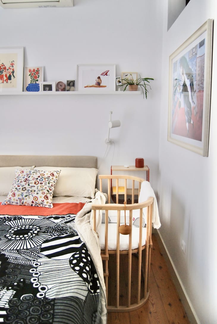 Primary bedroom with white walls, white floating shelves with art leaning on them, and a black and white marimekko blanket on top of the bed and a small oval natural wood spindle bassinet is by the bed.