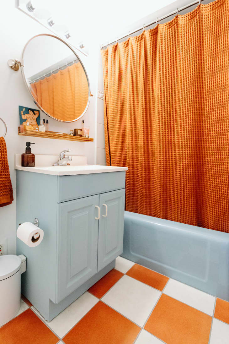 Round mirror hangs on wall above sink with blue cabinet in bathroom with orange accents.