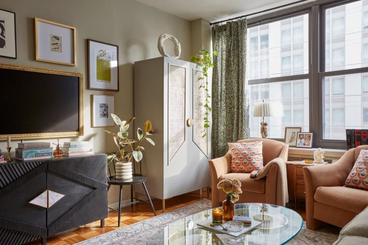 Living room with sage/olive colored walls, gallery wall, black credenza, 2 peach armchairs with throw pillows, gray patterned rug, gray wardrobe/cabinet, patterned floor to ceiling curtains, sofa with patterned throw pillows, glass and gold oval coffee table