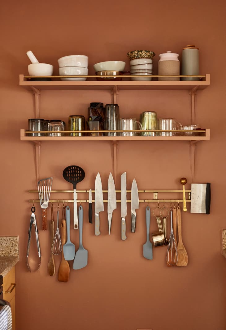 Racks on terra cotta colored kitchen wall with small bowls, vessels, mugs, knives, utensils