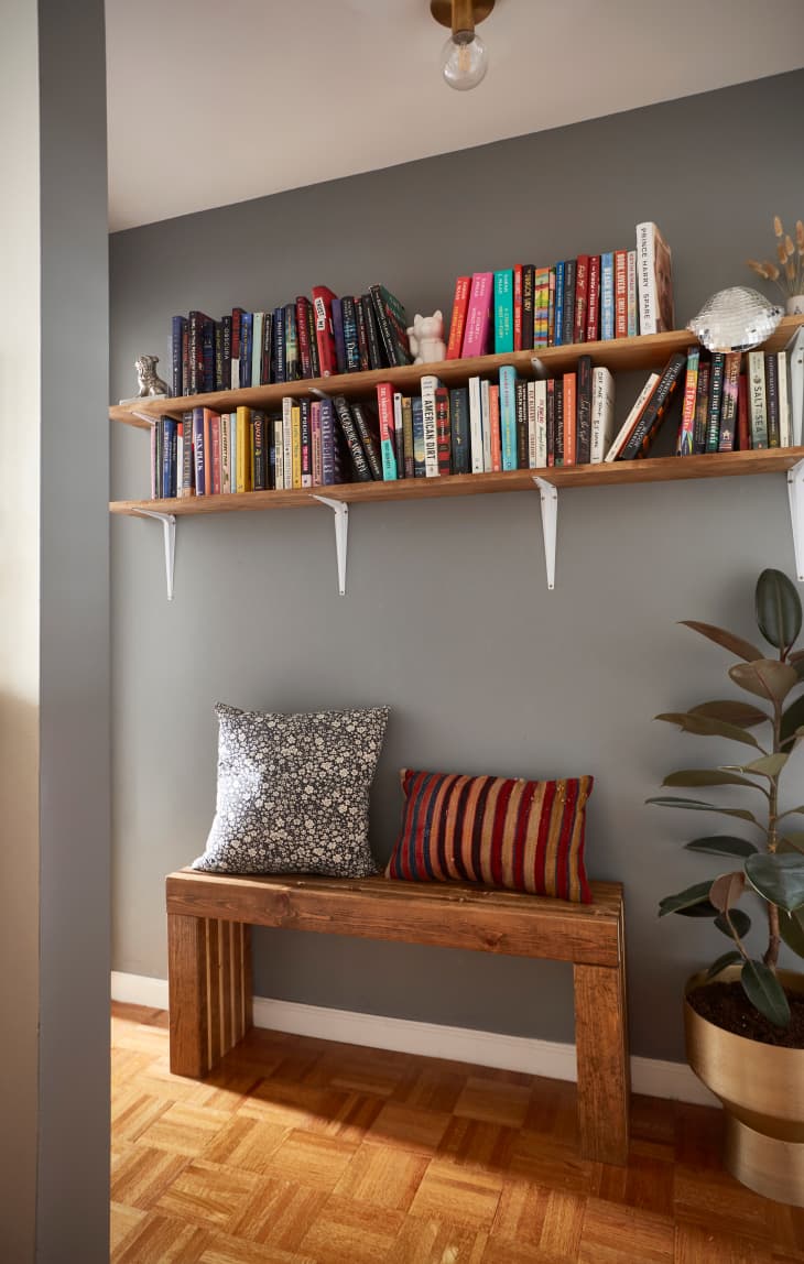 hallway area with a wood bench with pillows, a rubber plant on the floor in a gold planter, 2 shelves above with books, parquet wood floor