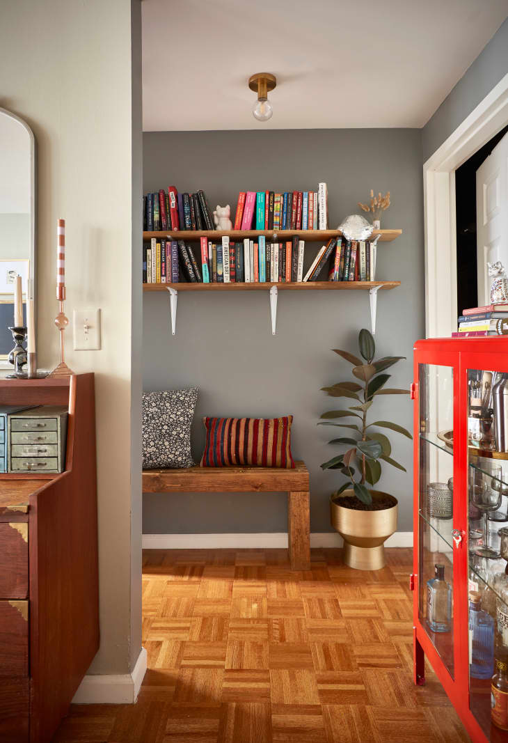 view of hallway area with a wood bench with pillows, a rubber plant on the floor in a gold planter, 2 shelves above with books, parquet wood floor