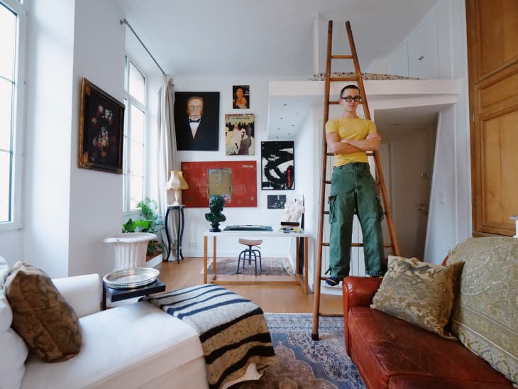 damon d standing in living room. White walls, wood ladder up to loft, large paintings, rust leather sofa, art objects