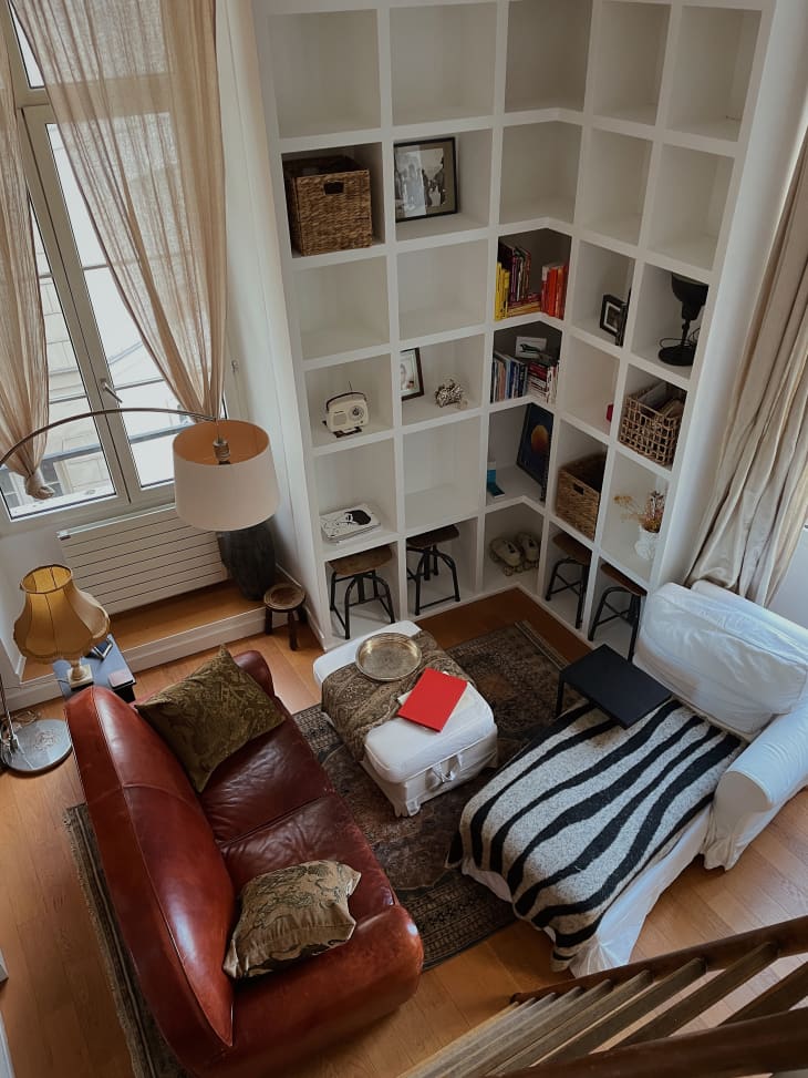 View of living room from loft above. Floor to ceiling white bookshelves, large windows, rust/deep red leather sofa, white chaise with black and white throw