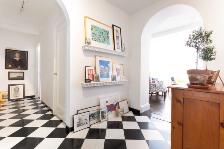 hallway area with black and white checkered floor, arched doorway, wood dresser, art on built-in shelves. Living room visible through arch on right, entry way/front door visible through arch on left