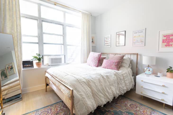 Bedroom in studio apartment with gold framed bed made with white linen, pink pillow and a white nightstand beside.