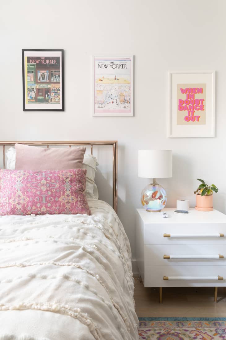 Bedroom in studio apartment with gold framed bed made with white linen, pink decorative pillows and a white nightstand beside the bed. Posters decorate the wall.