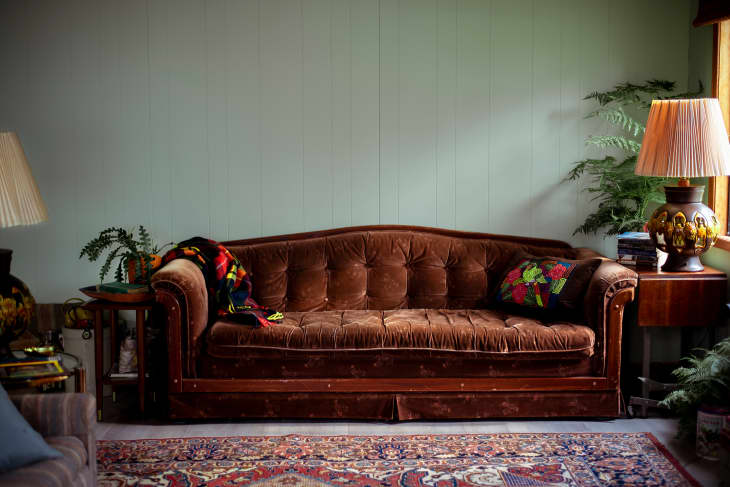 brown velvet sofa with black red and green pillow, throw. Vintage lamp and wood side table, pale green wall