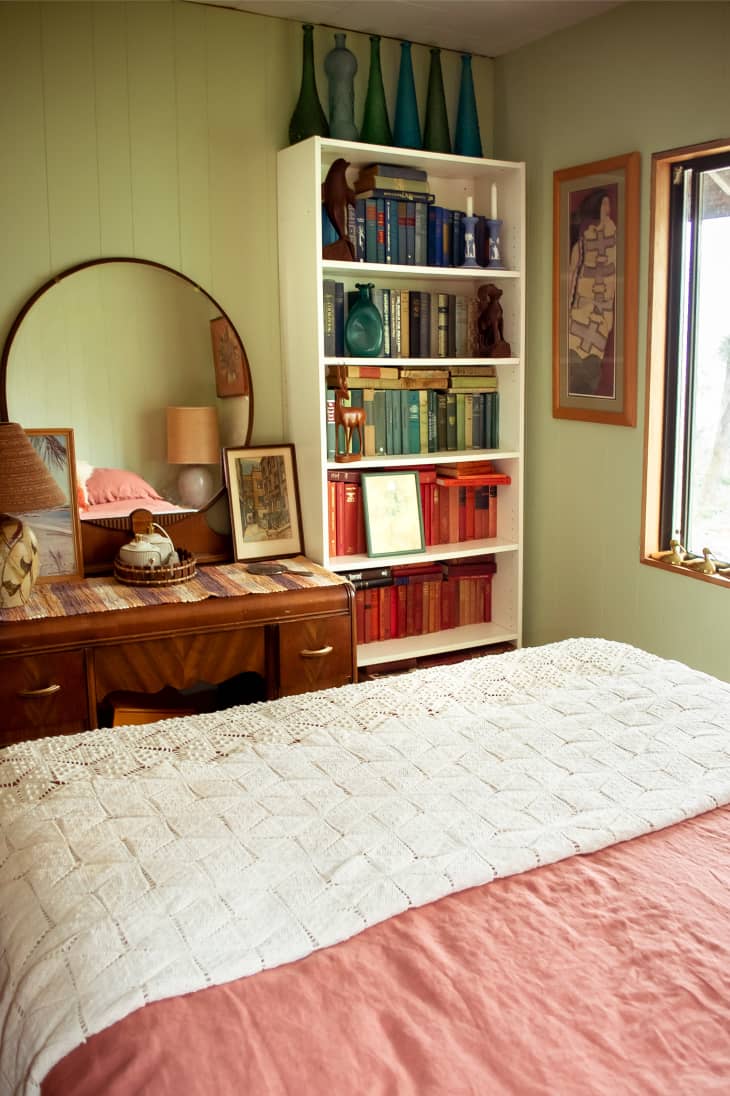 wall of guest room, part of bed is visible. Bookshelf, deco vanity table with round mirror