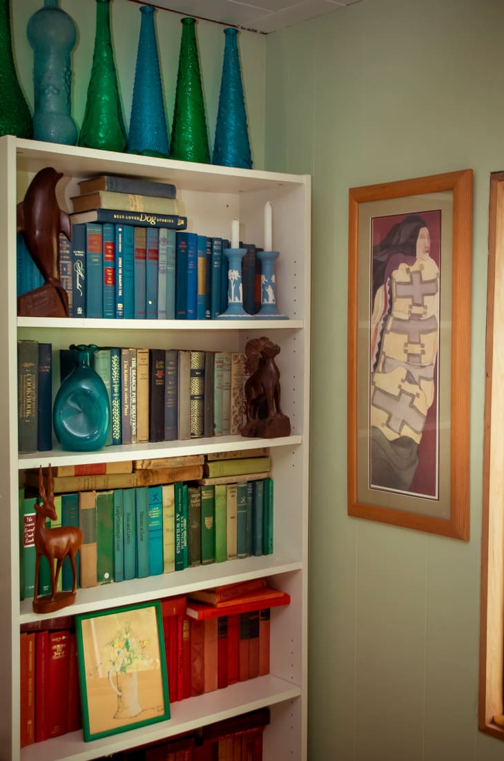 detail of bookshelf with color-arranged books. pale green walls, blue and green vases on top of shelves, framed painting on wall