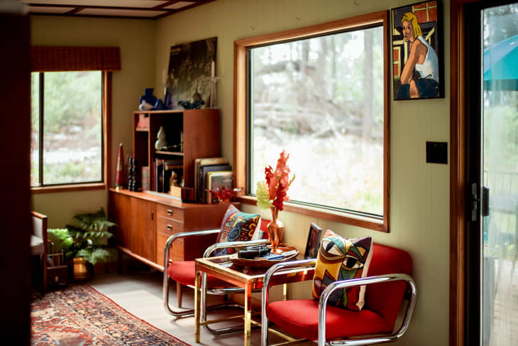 corner of living room with 2 red chairs with metal arms/legs, small table between them. Decorative throw pillows. There is a wood credenza with shelves above. Books and objets on it. Window shows lake view