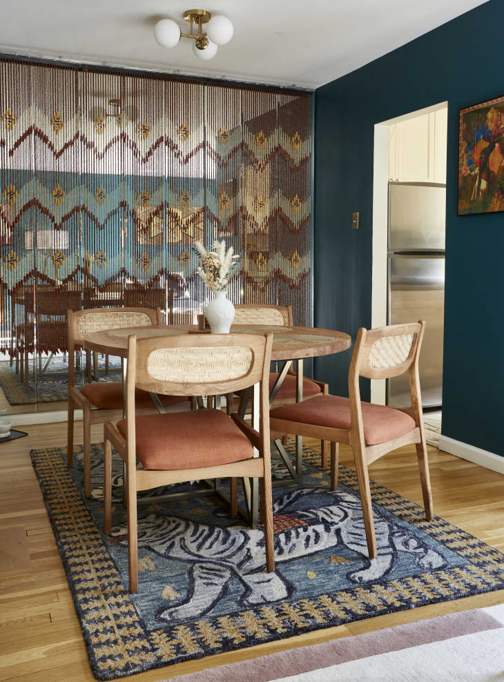 Dining room with mirror wall. beads hanging in front of mirror to create pattern. Round wood dining table with rattan and pale orange fabric chairs. One teal wall on the right. Area rug with tigers