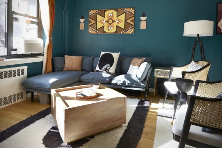 Living room with dark teal wall, artwork, gray sofa, wood block coffee table, rattan and fabric accent chairs
