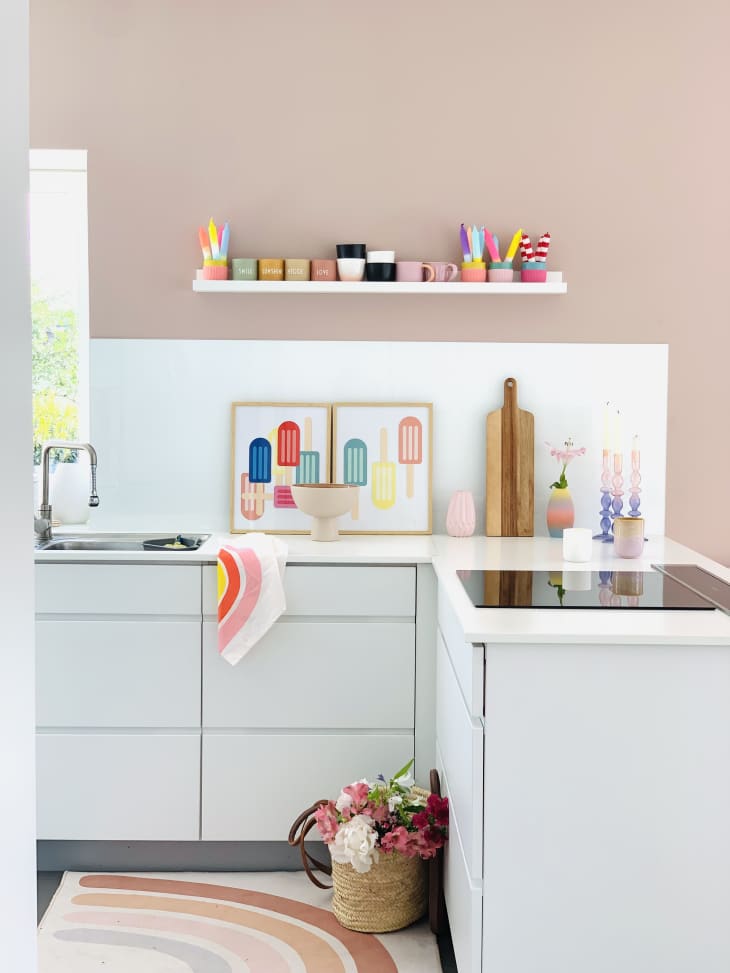 white cabinet, electric stove, rainbow rug, floating shelf, colorful cups, pastel pink wall, cutting board