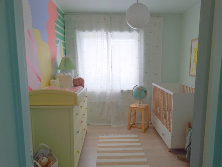 multi color splat mural with stripes throughout, white and yellow striped runner, wood floors, globe, yellow stool, changing table, yellow dresser, white orb lighting fixture, white crib with wood bars, light and airy curtains, basket of toys